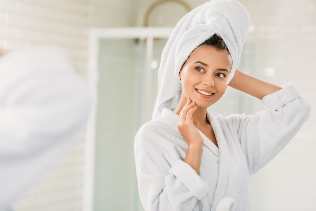 A woman is standing in her bathroom with her hair in a towel and dressed in a robe as she smiles at herself in the mirror.